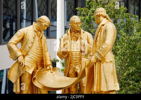 Diese vergoldete Bronzestatue, bekannt als die ‘Golden Boys’, befindet sich vor der New Birmingham Library und dem ICC in der Broad Street, Birmingham. Die Statue wurde 2022 nach der Renovierung des hundertjährigen Platzes an diesen neuen Ort verlegt. Die Statue ehrt Matthew Boulton, James Watt und William Murdoch. Sie werden gezeigt, wie sie die Pläne der Dampflokomotive untersuchen. Zusammen revolutionierten diese drei die Dampfmaschine – die Technologie, die buchstäblich die industrielle Revolution vorantreiben würde. Stockfoto