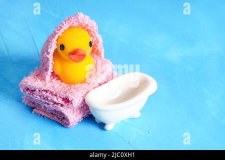 Gelbe Babyente in ein Handtuch gewickelt, das von einer weißen Miniaturbadewanne auf einem hellblauen Hintergrund mit Platz zum Kopieren auf einen Stapel weicher Handtücher gelegt wird. Kreativ Stockfoto
