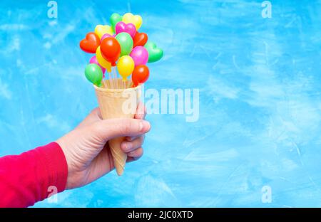 Waffelkegel mit kleinen herzförmigen Dekorationsballons in der Hand auf blauem Hintergrund mit Kopierraum. Eis-Sommerparty-Konzept. Stockfoto