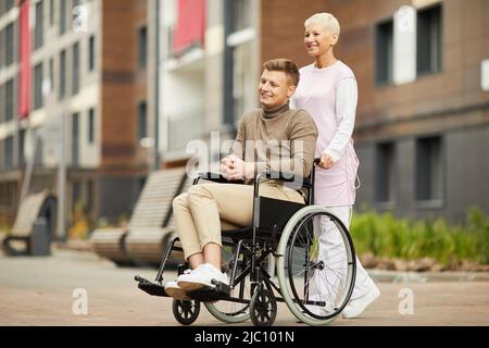 Positiv schöner junger Mann in lässigem Outfit, der im medizinischen Zentrum rehabilitieren muss, Krankenschwester, die den Rollstuhl mit dem jungen Mann schiebt Stockfoto