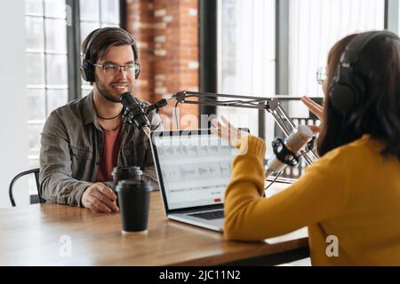 Lächelnder, gutaussehender Mann, der mit einer gestikulierenden, hübschen Radiomoderatorin spricht, während sie im Studio Live-Audio-Podcast aufnehmen. Zwei Personen, die einen Laptop, Kopfhörer und Audiogeräte für die Erstellung eines Podcasts verwenden Stockfoto