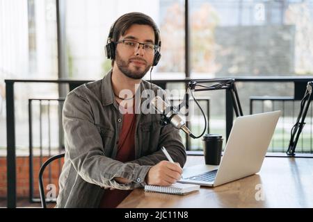 Portrait von fröhlichen jungen Mann Host Aufnahme Podcast im Studio. Ein gutaussehender lächelnder Kerl mit Kopfhörern und Brillen, der am Tisch mit Laptop und Mikrofon sitzt, die Kamera anschaut, Stift und Notebook benutzt Stockfoto