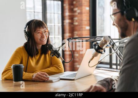 Junge schöne Frau Radio Gastgeber lächelnd, während Mann Gast im Studio interviewen, während Podcast für Online-Show aufnehmen. Zwei Podcasts in Kopfhörern, die sich gegenseitig anguckend und Audio-Podcasts aufzeichnend sind Stockfoto