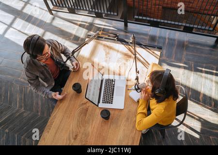 Draufsicht auf zwei Personen in Kopfhörern, die Audio-Podcast mit Mikrofonen im Sendestudio aufnehmen. Junge Frau im Gespräch mit ihrem männlichen Kollegen, während sie Podcast aufzeichnen. Live-Streaming Stockfoto