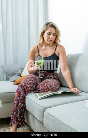 Junge blonde Frau, die auf dem Sofa sitzt und einen Gemüse-Smoothie trinkt Stockfoto