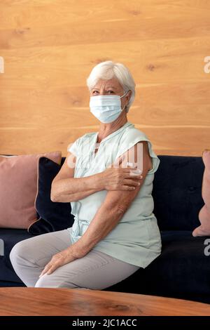 Porträt einer älteren kaukasischen Frau mit Gesichtsmaske, die ihren Arm mit Band hält und zu Hause sitzt Stockfoto