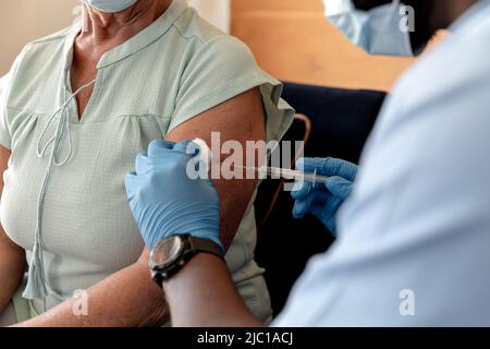 Mittlerer Abschnitt eines afroamerikanischen männlichen Gesundheitarbeiters, der einer kaukasischen älteren Frau eine Injektion verabgab Stockfoto