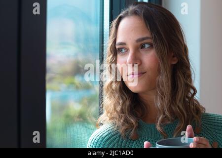 Kaukasische junge Frau mit einer Kaffeetasse und Blick aus dem Fenster zu Hause Stockfoto