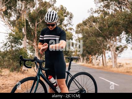 Professionelle Radfahrerin, die mit ihrem Fahrrad auf einer leeren Straße steht und Fitness-Tracker überprüft Stockfoto