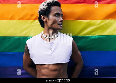 Porträt eines hübschen Mannes, der wegschaut, während er gegen die LGBT-Flagge des Regenbogens steht Stockfoto