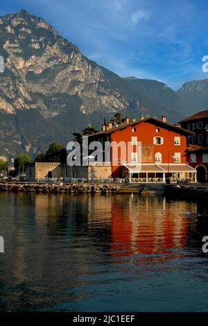 Die Casa Beust, ihre roten ockerfarbenen Wände spiegeln sich in der ruhigen Sonne des Gardasees am frühen Morgen des Julis wider, wurde 1400s als venezianische Villa am Ufer von Torbole sul Garda, Trentino-Südtirol, Italien, erbaut. Seitdem diente es als Treffpunkt für Künstler und beherbergt heute ein Restaurant, eine Pizzeria, ein Café und Apartments. Stockfoto