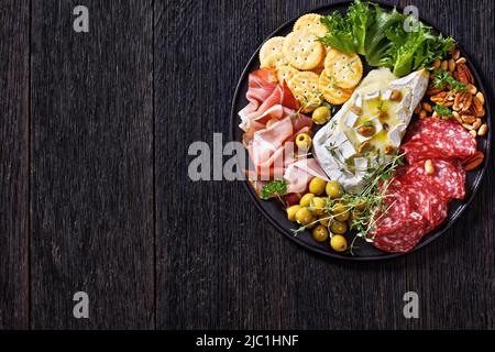 Herzhaft gebackener Brie mit Crackern, spanischem Salchichon, Salami-Würsten, dünn geschnittenem geräuchertem Schinken, grünen Oliven, Salat, Erdnüssen, Pecan auf schwarzer Platte Stockfoto