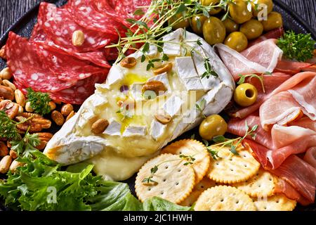 Herzhaft gebackener Brie mit Crackern, spanischer Salchichon, Salami-Würste, dünn geschnittener Räucherschinken, grüne Oliven, Salat, Erdnüsse, Pekannüsse auf schwarzem Teller, Stockfoto