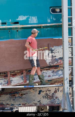 Ein Mann, der auf einer Legierungsplanke steht und mit einer Walze die Seite eines großen Fischtrawlers auf einem Slipway malt Stockfoto