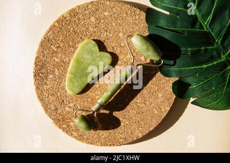 Jade Gua Sha Scraper und Face Roller Massagegerät auf einem runden Korkständer mit Monstera-Blatt. Hartes Licht, Schatten, das Konzept der Selbstversorgung. Gesichtspflege. Zer Stockfoto
