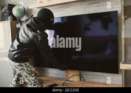 Ein maskierter Dieb entfernt einen teuren Fernseher von der Wand. Das Konzept des Wohnungsdiebstahls Stockfoto