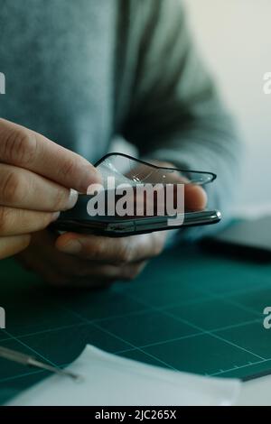 Ein junger Kaukasusmann entfernt den zerbrochenen Bildschirmschutz eines Telefons und sitzt an einem Tisch Stockfoto