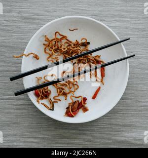 Hochwinkelansicht eines weißen Tellers mit den Resten von Chicken Yakisoba Nudeln und einem Paar Essstäbchen, auf einem Tisch mit einem grauen Tischset Stockfoto