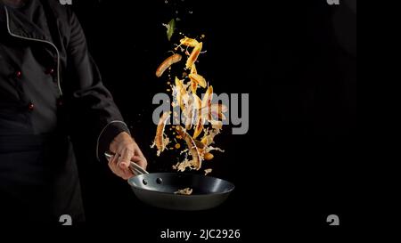 Anonymer Koch in Uniform wirft appetitlich gebratenes Schupfnudeln mit Würstchen in der Bratpfanne vor schwarzem Hintergrund während des Kochvorgangs im Studio Stockfoto