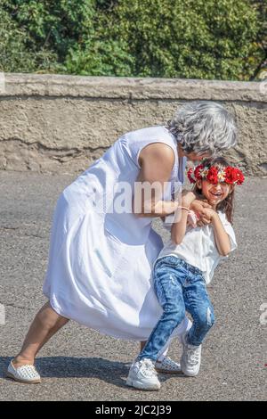 7 15 2019 Tiflis Georgia Großmutter in weißem Kleid tanzt mit einer jungen Enkelin in Blumenschmuck im Freien, die sie taucht und wirbelt Stockfoto