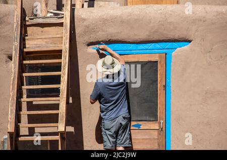 Zurück der gebürtigen amerikanischen Mann in Shorts und T-Shirt und Sonnenhut Malerei hellblau-türkis rund um die Tür eines Schlamm adobe Pueblo mit Holztreppe Stockfoto