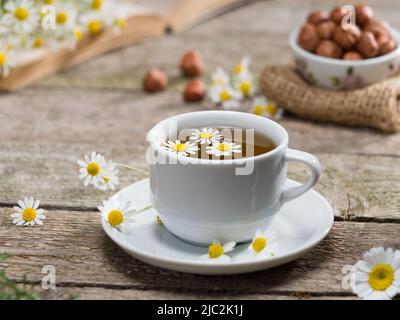 Gesunder Kamillentee und Kamillenkräuter auf Holztisch. Bücher und Nüsse im Hintergrund. Entspannendes Kräutertees-Konzept. Stockfoto