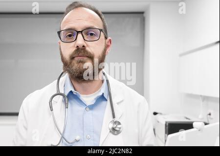 Arzt im mittleren Alter, der in der Klinik einen weißen medizinischen Mantel mit ernstem Ausdruck trägt. Hochwertige Fotos Stockfoto