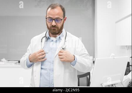 Arzt im mittleren Alter, der in der Klinik einen weißen medizinischen Mantel mit ernstem Ausdruck trägt. Hochwertige Fotos Stockfoto