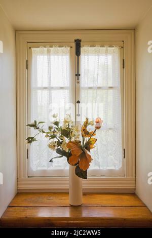 Weiße Keramikvase mit Seidenblumen auf Fensterbank im Hauptschlafzimmer im Obergeschoss im alten rekonstruierten 1850s-Blockhaus. Stockfoto