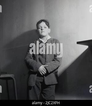 1950s, historisches Bild eines Schuljungen, der einen Pullover und eine Jacke trägt, mit dem Rücken gegen eine Wand im Klassenzimmer steht und die Kamera anschaut, England, Großbritannien. Stockfoto