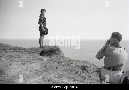 1938, historisch, August, Vorkriegszeit II und am Rand einer Klippe, ein Pfadfindermeister, der ein Foto eines Pfadfinderjungen macht, der aufrecht steht, seinen Pfadfinderhut hält und über den Ozean blickt, als Andenken an das Pfadfinderlager auf der Isle of Wright, Südengland, Großbritannien. Stockfoto