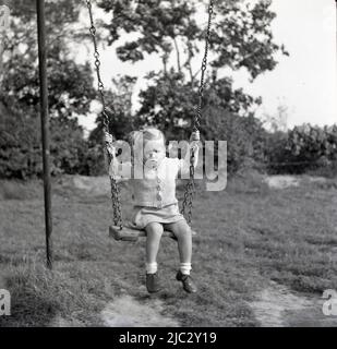 1950s, historisch, draußen auf einem Spielplatz auf dem Land, ein kleines Mädchen, das auf dem hölzernen Sitz einer Schaukel der Zeit sitzt und an den Metallketten festhält, England, Großbritannien. Stockfoto