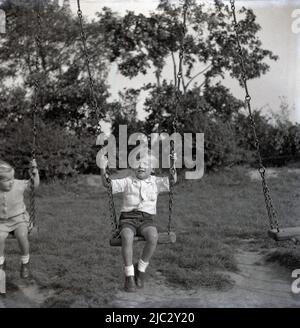 1950s, historisch, draußen auf einem Spielplatz auf dem Land, ein kleiner Junge, der auf dem hölzernen Sitz einer Schaukel der Zeit sitzt und die Metallketten hält, England, Großbritannien. Stockfoto