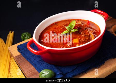 Food-Konzept hausgemachte italienische Bolognese-Sauce in roten Ofenaufläufen auf Holzbrett mit schwarzem Kopierraum Stockfoto