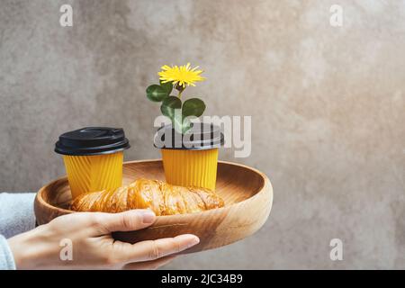 Zwei gelbe Pappbecher für Kaffee oder Tee und frische Croissants auf einem Holztablett. Bestellen Sie die Bestellung in den Händen der Frauen. Dandelion Blume mit Kleeblättern für romantisches Frühstück am St. Patrick's Day Copy Space Stockfoto