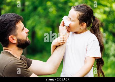 Seitenansicht des bärtig-hübschen Mannes im mittleren Alter, Vater, der mit einem weißen Taschentuch die Nase seiner Töchter abwischt und im Wald spaziert. Stockfoto