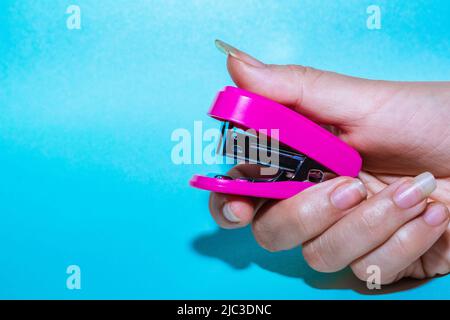 Junge lateinische Frauenhand, mit langen Fingernägeln, die einen kleinen rosafarbenen Hefter halten, der ein Blatt Papier heften soll, auf blauem Hintergrund. Gegenstand für aus Stockfoto