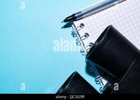 Detail von Fernglas, Notizbuch und schwarzem Stift auf blauem Hintergrund. Konzept von Reisen, Vogelsuche und Sichtung. Kleines Notizbuch mit leeren Seiten. Stockfoto