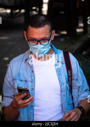 Medellin, Antioquia, Kolumbien - Februar 19 2022: Südamerikanischer junger Mann mit blauem Hemd und Gesichtsmaske sieht sein Telefon aus Stockfoto