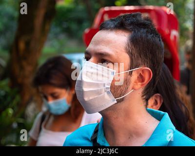 Medellin, Antioquia, Kolumbien - Februar 19 2022: Südamerikanischer Mann benutzt eine weiße Maske in der Straße in der Nähe anderer Menschen Stockfoto