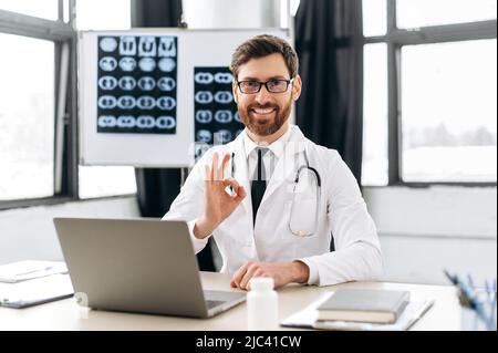 Positiv selbstbewusster kaukasischer Arzt in einer medizinischen Uniform und mit Brille, sitzt an einem Schreibtisch in seinem medizinischen Büro, schaut auf die Kamera, zeigt ein Zeichen der Zustimmung, OKAY Geste, lächelt freundlich Stockfoto