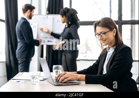 Die smarte kaukasische Frau mit Brille, im Business-Anzug, sitzt am Arbeitsplatz im Büro, arbeitet mit einem Laptop an einem neuen Projekt und vor dem Hintergrund diskutieren ihre Kollegen über die Finanzstrategie Stockfoto