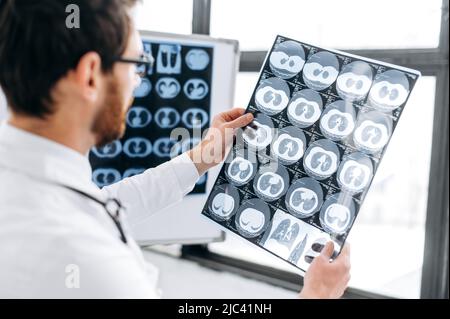 Röntgenuntersuchung. Fokussierter intelligenter Arzt, der Röntgenaufnahmen in seinem Kabinett ansah, kaukasischer Therapeut in weißer medizinischer Uniform und Brille konzentrierte die Röntgenaufnahmen des lernenden Patienten, der in seiner Arztpraxis stand Stockfoto