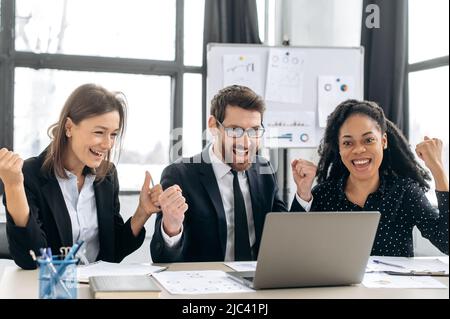 Fröhliches kreatives Geschäftsteam, das zusammen arbeitet. Kollegen aus verschiedenen Rassen denken gemeinsam an Strategien, sitzen im modernen Büro, nutzen Laptops, bekommen gute Angebote, profitieren, lächeln, Geste der Hände Stockfoto