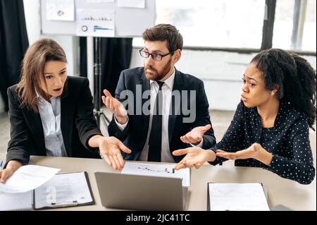 Verblüfft überraschten die Kollegen, verwirrt von den Informationen, die sie sahen, über einen Verlust der Ergebnisse des Gewinns aus dem Projekt, erlebten negative Emotionen, sprachen am Schreibtisch im Büro Stockfoto