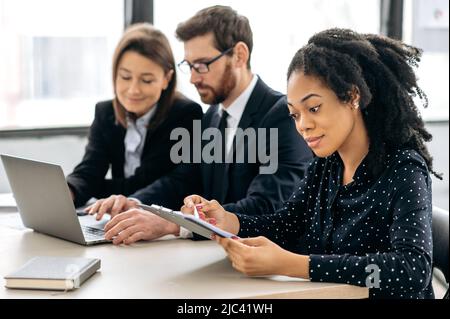 Fokussierte, intelligente afroamerikanische junge Frau im Hemd, eine Finanzmanagerin oder ein ceo, die mit ihren Kollegen im Büro an einem Schreibtisch sitzt, untersucht konzentriert die Dokumente, den Finanzbericht Stockfoto