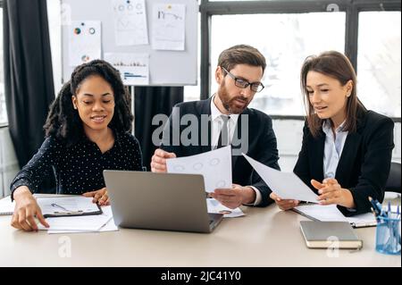 Multirassische kreative Kollegen führen gemeinsam Brainstorming zu einem neuen Start-up durch, sitzen im modernen Büro, studieren Dokumente, nutzen Laptops und diskutieren den Finanzgeschäftsplan. Konzept der Teamzusammenarbeit Stockfoto