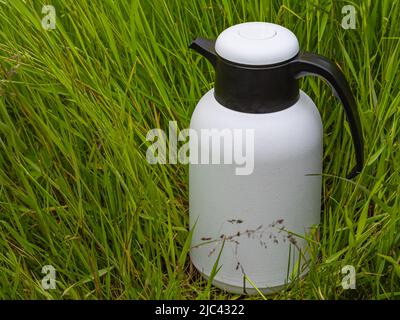 Weiße Thermoflasche isoliert auf grünem Gras. Ein Heißgetränk duht auf einem Rasen im Park. Niemand, selektiver Fokus, Platz für Text kopieren Stockfoto