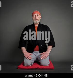 Kopf- und Schulterporträt eines kahlen und bärtigen älteren Mannes, der einen kurzen Kimono trägt und in einer traditionellen japanischen Seiza-Position auf einem Kissen pa sitzt Stockfoto