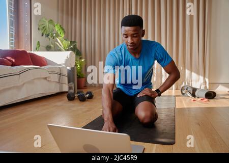 Junger afroamerikanischer schwarzer Mann, der gerade dabei ist, mit seinem Laptop an einem Online-Trainingskurs teilzunehmen, auf einer Yogamatte zu sitzen und von zu Hause aus zu trainieren Stockfoto